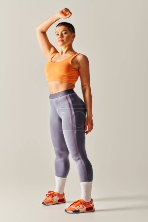 Körperbewusstsein, athletische und kurzhaarige Frau, die auf grauem Hintergrund posiert, kurviges Fitnessmodel, stehend mit erhobener Hand, Ausdauer und Empowerment, Generation z, volle Länge 