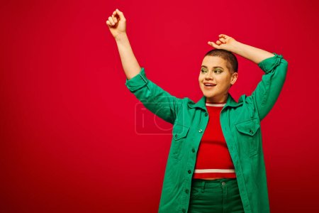 Mode und Stil, fröhliche und kurzhaarige Frau im grünen Outfit posiert mit erhobenen Händen auf rotem Hintergrund, Generation z, Jugendkultur, moderner Hintergrund, Individualität, persönlicher Stil 