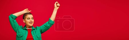 Mode und Stil, aufgeregte und kurzhaarige Frau im grünen Outfit posiert mit erhobenen Händen auf rotem Hintergrund, Generation z, Jugendkultur, moderner Hintergrund, Individualität, persönlicher Stil, Banner 