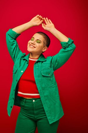 Mode und Stil, fröhliche und kurzhaarige Frau im grünen Outfit posiert mit erhobenen Händen auf rotem Hintergrund, lächelnd, Generation z, Jugendkultur, moderner Hintergrund, Individualität, persönlicher Stil 