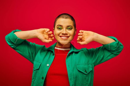 fashion statement, cheerful and short haired woman in green outfit posing on red background, generation z, youth culture, modern backdrop, individuality, personal style, looking at camera