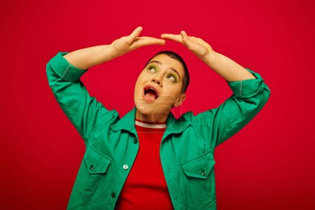 fashion and style, amazed and short haired woman in green outfit posing with raised hands on red background, looking up generation z, youth culture, vibrant backdrop, individuality, personal style 