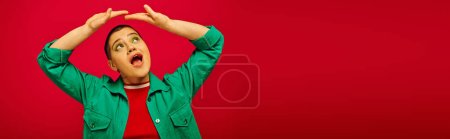 Mode und Stil, staunende und kurzhaarige Frau im grünen Outfit posiert mit erhobenen Händen auf rotem Hintergrund, schaut nach oben Generation z, Jugendkultur, lebendige Kulisse, persönlicher Stil, Banner 