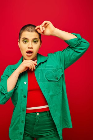 Mode und Stil, schockierte und kurzhaarige Frau in grünem Outfit posiert mit händennahem Gesicht auf rotem Hintergrund, blickt in die Kamera, Generation Z, Jugendkultur, lebendige Kulisse, Individualität 