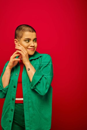 fashion and trends, tattooed, cheerful and short haired woman in green outfit posing on red background, looking away, generation z, youth culture, vibrant backdrop, individuality 