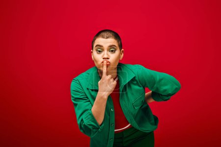 Photo for Fashion and style, tattooed, short haired woman in green outfit posing with finger near lips on red background, looking at camera, generation z, youth culture, vibrant backdrop, individuality - Royalty Free Image