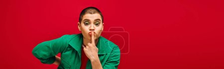 Photo for Fashion and style, tattooed, short haired woman in green outfit posing with finger near lips on red background, looking at camera, generation z, youth culture, vibrant backdrop, individuality, banner - Royalty Free Image