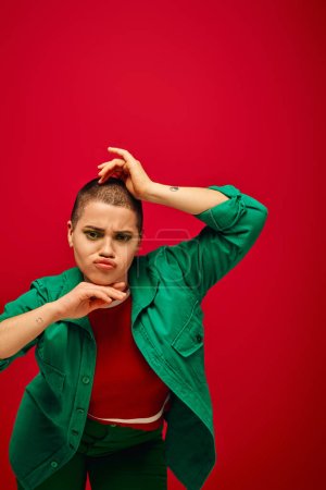 fashion and style, emotional and tattooed, short haired woman in green outfit pouting lips on red background, looking at camera, generation z, youth culture, vibrant backdrop, individuality 