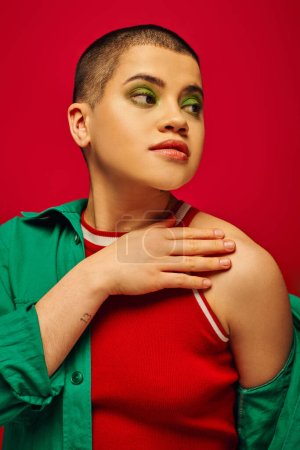 Photo for Fashion trend, youthful and tattooed, short haired woman in green outfit posing on red background, looking away, generation z, youth, vibrant backdrop, bold makeup, personal style, portrait - Royalty Free Image