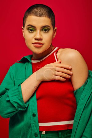 Modetrend, jugendlich und tätowiert, kurzhaarige Frau in grünem Outfit posiert auf rotem Hintergrund, blickt in die Kamera, Generation Z, Jugend, lebendige Kulisse, fettes Make-up, persönlicher Stil, Porträt 