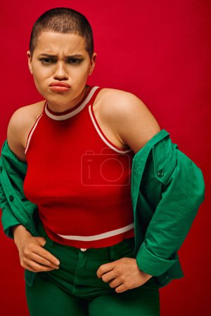 Photo for Fashion trend, emotional and tattooed, short haired woman in green outfit posing on red background, generation z, youth, pouting lips, vibrant backdrop, bold makeup, personal style, portrait - Royalty Free Image