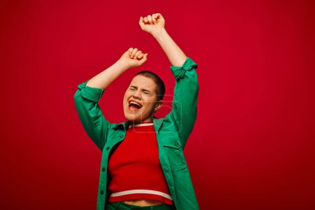 Photo for Fashion statement, emotional and tattooed, short haired woman in green outfit singing on red background, generation z, raised hands, youth, vibrant backdrop, bold makeup, personal style - Royalty Free Image