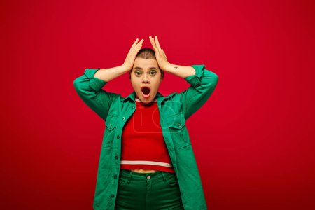 fashion trend, shocked and tattooed, short haired woman in green outfit touching head on red background, looking away, generation z, youth, vibrant backdrop, individuality, personal style 