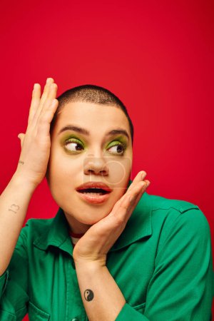 fashion choices, amazed and tattooed, short haired woman in green outfit touching head on red background, looking away, generation z, youth, vibrant backdrop, individuality, personal style 