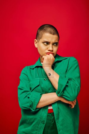 Photo for Fashion choices, displeased and tattooed, short haired woman in green outfit touching chin on red background, looking away, generation z, youth, vibrant backdrop, emotional and youthful - Royalty Free Image