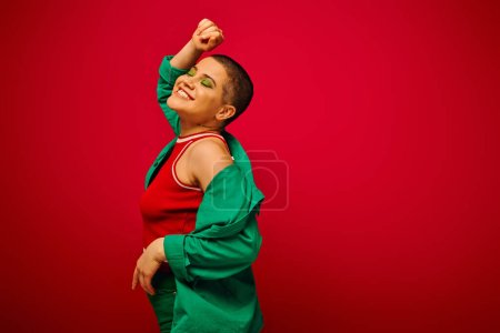 fashion statement, bold makeup, satisfied and tattooed, short haired woman in green outfit smiling on red background, generation z, youth, vibrant backdrop, individuality, personal style 