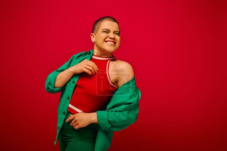 fashion statement, bold makeup, pleased and tattooed, short haired woman in green outfit smiling on red background, generation z, youth, vibrant backdrop, individuality, personal style 