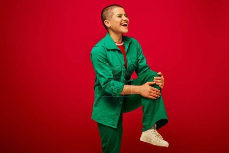 stylish look, bold makeup, cheerful and tattooed, short haired woman in green outfit posing on red background, looking away, generation z, youth, vibrant backdrop, individuality, personal style 