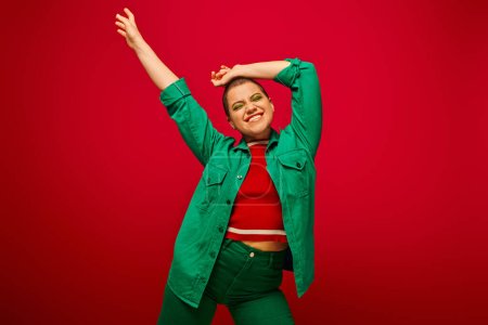 stylish outfit, bold makeup, cheerful and tattooed, short haired woman in green outfit posing on red background, generation z, youth culture, vibrant backdrop, personal style 