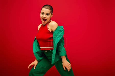 stylish appearance, bold makeup, amazed and tattooed, short haired woman in green outfit posing on red background, generation z, youth, vibrant backdrop, individuality, personal style 