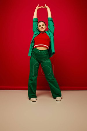 fashion statement, vibrant backdrop, cheerful and young woman in stylish attire posing with raised hands on red background, full length, generation z, youth culture, personal style, curvy fashion 