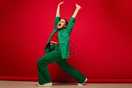 Mode-Statement, lebendige Kulisse, aufgeregte und junge Frau in stilvoller Kleidung posiert mit erhobenen Händen auf rotem Hintergrund, volle Länge, Generation z, Jugendkultur, persönlicher Stil, kurvige Mode 