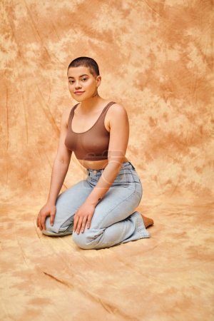 Photo for Representation of body, acceptance, curvy young and tattooed woman in jeans and crop top sitting on mottled beige background, personal style, self-acceptance, generation z, denim fashion, tattooed - Royalty Free Image