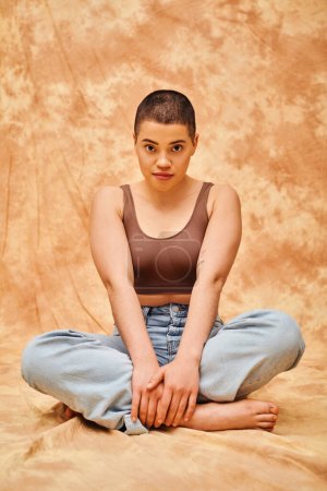 representation of body, casual wear, curvy, young and tattooed woman in jeans and crop top sitting on mottled beige background, personal style, self-acceptance, generation z, denim fashion, tattooed 