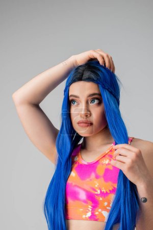 self expression, young woman adjusting blue hair and looking away on grey background, isolated, fashion choices, stylish look, colorful clothes, casual attire, generation z fashion, long hair