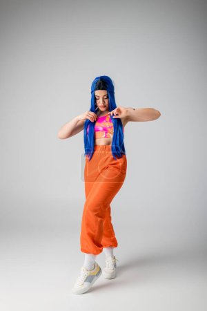 tendencias de moda, mujer joven tatuada con el pelo teñido de azul posando en ropa de colores sobre fondo gris, longitud completa, aspecto funky, individualismo, estilo moderno, moda urbana, color vibrante, modelo 