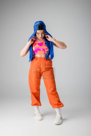 Foto de Modelo femenino, mujer joven tatuada con el pelo azul posando en ropa colorida sobre fondo gris, longitud completa, aspecto funky, individualismo, estilo moderno, moda urbana, color vibrante - Imagen libre de derechos