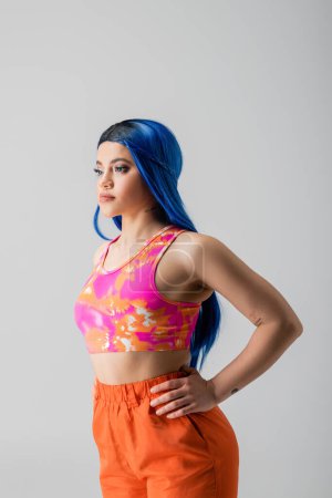 Photo for Fashion forward, tattooed young woman with blue hair posing with hands on hips in colorful clothes on grey background, individualism, modern style, urban fashion, vibrant color, female model - Royalty Free Image
