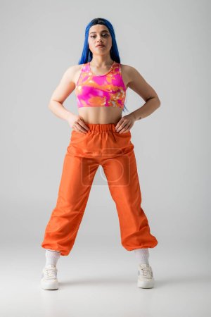 Photo for Fashion, tattooed young woman with blue hair posing with hands on hips and standing in colorful clothes on grey background, full length, individualism, modern style, urban fashion, vibrant color - Royalty Free Image
