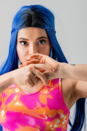 fashion forward, tattooed young woman with blue hair posing with hands near face on grey background, individualism, modern style, urban fashion, vibrant color, female model, looking at camera