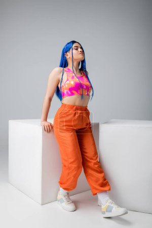 fashion trends, tattooed young woman with blue hair posing in colorful clothes near white cubes on grey background, full length, individualism, modern style, urban fashion, vibrant color, model 