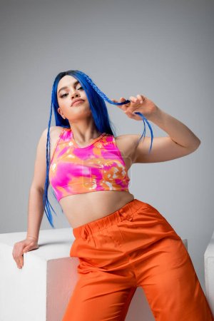 estilo rebelde, mujer joven tatuada con el pelo azul posando en ropa de colores cerca del cubo blanco sobre fondo gris, mirando a la cámara, individuo moderno, moda urbana, gen z