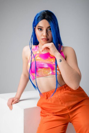 style rebelle, jeune femme tatouée aux cheveux bleus posant en vêtements colorés près du cube blanc sur fond gris, look élégant, regardant la caméra, individu moderne, mode urbaine, génération z 