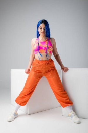 fashion forward, tattooed young woman with blue hair standing in colorful clothes near white cubes on grey background, full length, individualism, modern style, urban fashion, vibrant color, model 