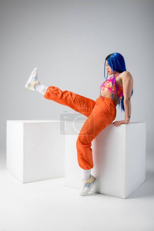 tendencias de moda, mujer joven tatuada con el pelo azul posando con la pierna levantada cerca de cubos blancos sobre fondo gris, longitud completa, individualismo, estilo moderno, moda urbana, color vibrante, modelo 
