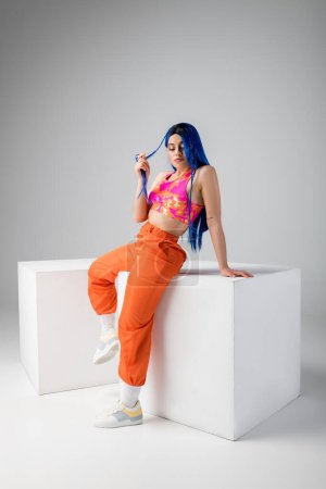 Photo for Fashion statement, tattooed young woman with blue hair posing in colorful clothes near white cubes on grey background, full length, individualism, modern style, vibrant color, urban fashion - Royalty Free Image