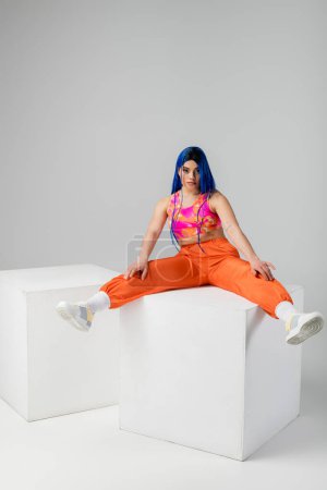 Foto de Tendencias de moda, mujer joven tatuada con el pelo azul sentado con las piernas extendidas en cubo blanco sobre fondo gris, longitud completa, individualismo, estilo moderno, moda urbana, color vibrante, modelo - Imagen libre de derechos