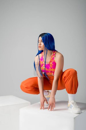 Mode und Stil, tätowierte junge Frau mit blauen Haaren sitzt auf einem weißen Würfel auf grauem Hintergrund, volle Länge, Individualismus, moderner Stil, urbane Mode, lebendige Farbe, Modell 
