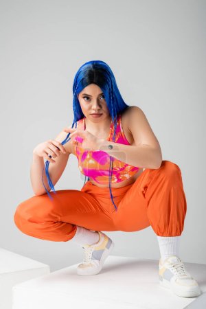 Foto de Cultura juvenil, mujer joven tatuada con el pelo azul sentado encima del cubo blanco sobre fondo gris, longitud completa, individualismo, estilo moderno, moda urbana, color vibrante, modelo - Imagen libre de derechos