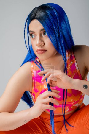 estilo rebelde, mujer joven tatuada con el pelo azul posando en ropa colorida aislada sobre fondo gris, aspecto elegante, mirando hacia otro lado, individuo moderno, moda urbana, generación z 