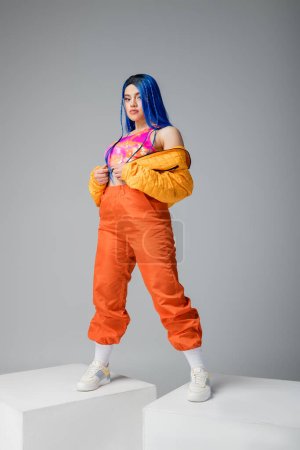 Photo for Full length, fashion forward, young woman with blue hair posing in colorful clothes on grey background, standing on white cubes, vibrant color, female model, individualism, urban fashion - Royalty Free Image