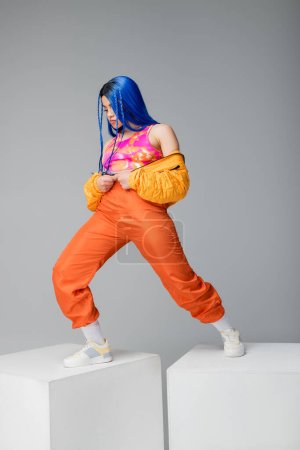 full length, fashion forward, young woman with blue hair posing in puffer jacket and orange pants on grey background, standing on white cubes, vibrant color, female model, urban fashion 