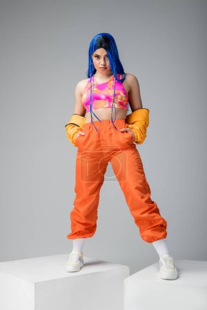 full length, fashion forward, tattooed woman with blue hair posing with hands in pockets of orange pants on grey background, standing on white cubes, vibrant color, female model, urban fashion 