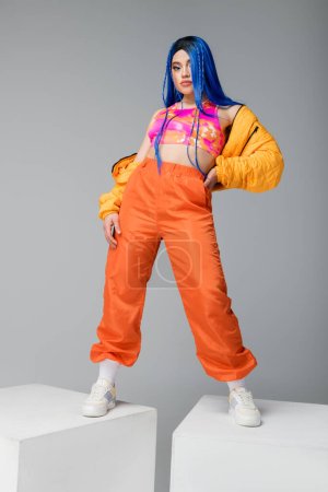 volle Länge, Mode nach vorne, weibliches Model mit blauen Haaren posiert in Pufferjacke und orangefarbener Hose auf grauem Hintergrund, mit der Hand an der Hüfte auf weißen Würfeln stehend, lebendige Farbe, urbane Mode 