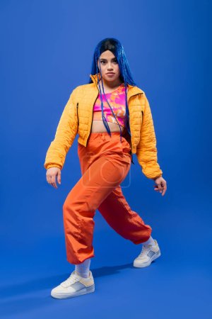 volle Länge, Mode nach vorne, junges weibliches Model mit blauen Haaren posiert in Pufferjacke und orangefarbener Hose auf blauem Hintergrund, lebendige Farbe, urbane Mode, Individualismus
