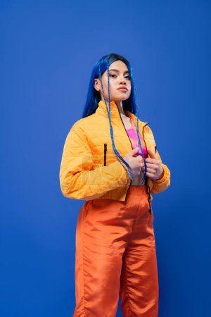 gefärbte Haare, Mode nach vorne, tätowiertes weibliches Model mit blauen Haaren posiert in Pufferjacke und orangefarbener Hose auf blauem Hintergrund, lebendige Farbe, urbane Mode, Individualismus, junge Frau 